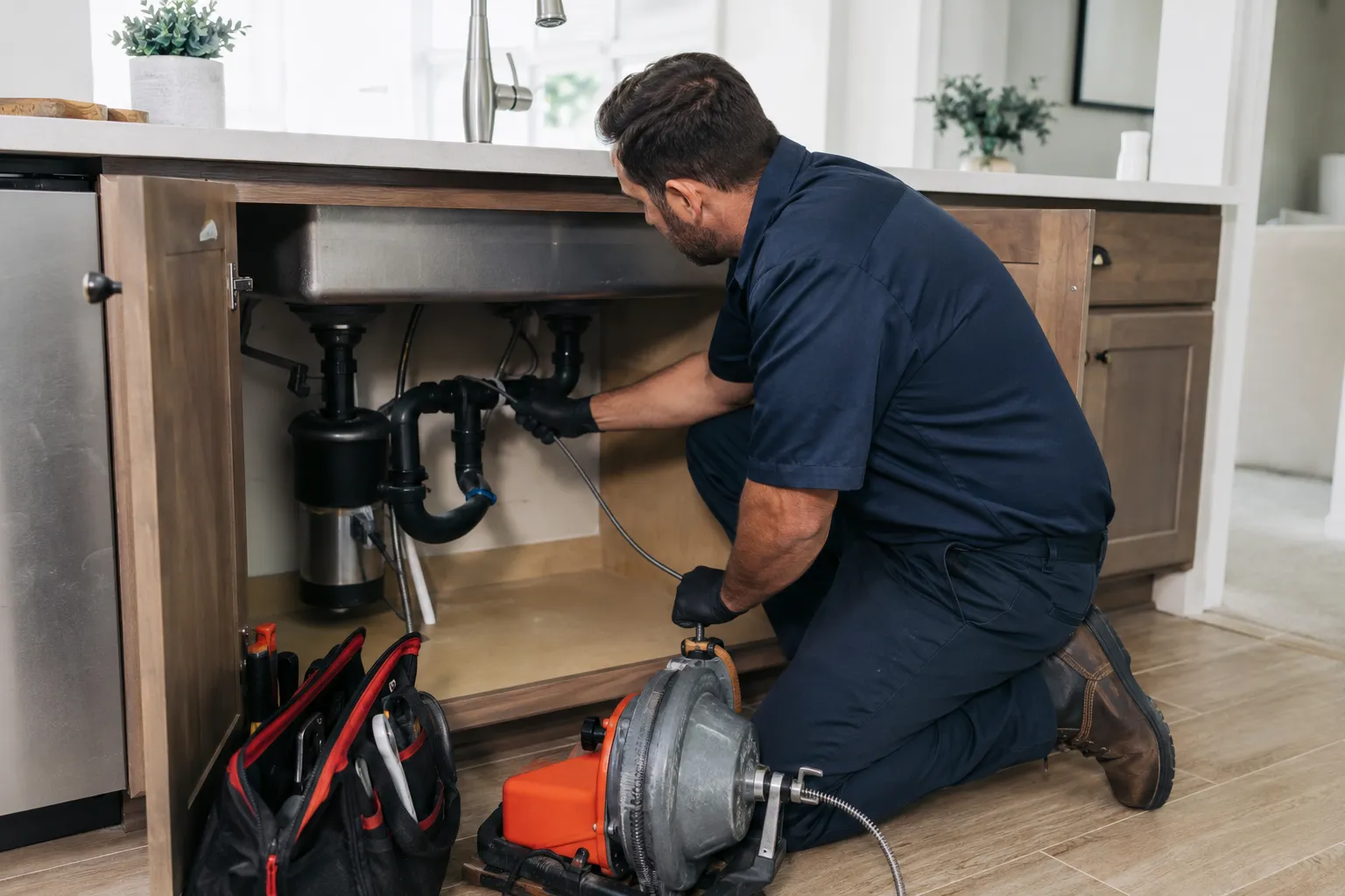 A technician working a drain machine cable into the trap under a kitchen sink